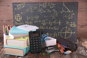 Des cahiers, livres et une calculatrice devant un tableau avec un exercice de math