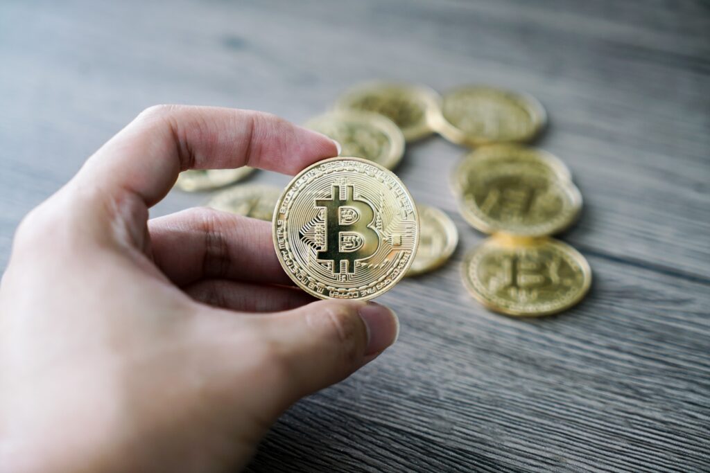 Une main tient une pièce de bitcoin dans la main avec en arrière plan des pièces de bitcoin sur une table