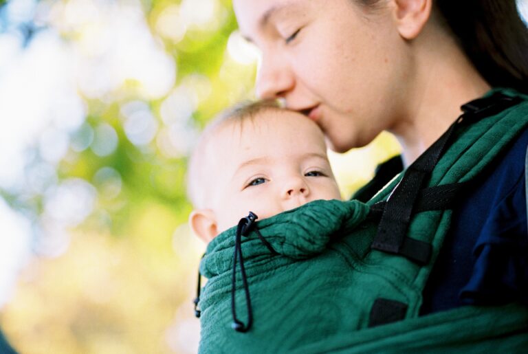 Maman faisant un bisou à son bébé assis dans le porte-bébé et souriant les yeux mi-clos